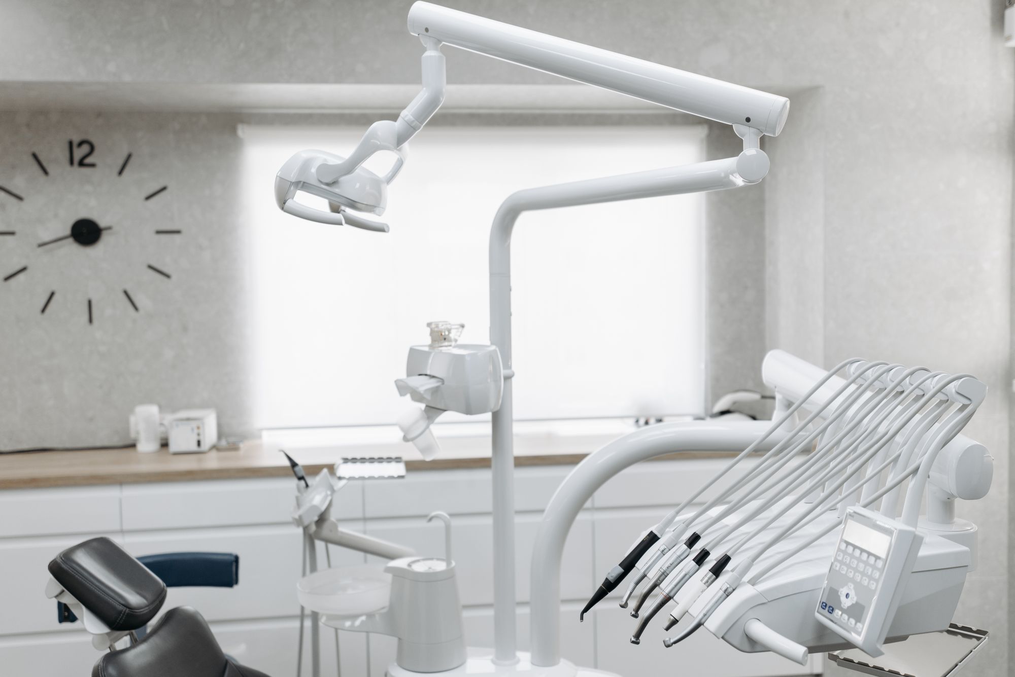 An empty dental treatment room with a patient chair, overhead lamp, and instrument tray beside a bright window.