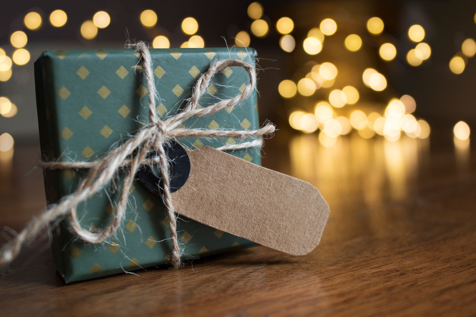A small gift box wrapped in green paper with twine and a blank tag, in front of warm string lights.