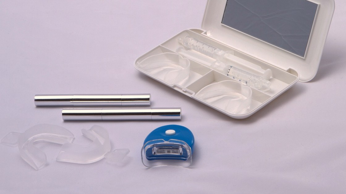 A take-home teeth whitening kit: a white case with a mirror holding clear custom trays and a gel syringe, beside two whitening gel pens and a small LED light.
