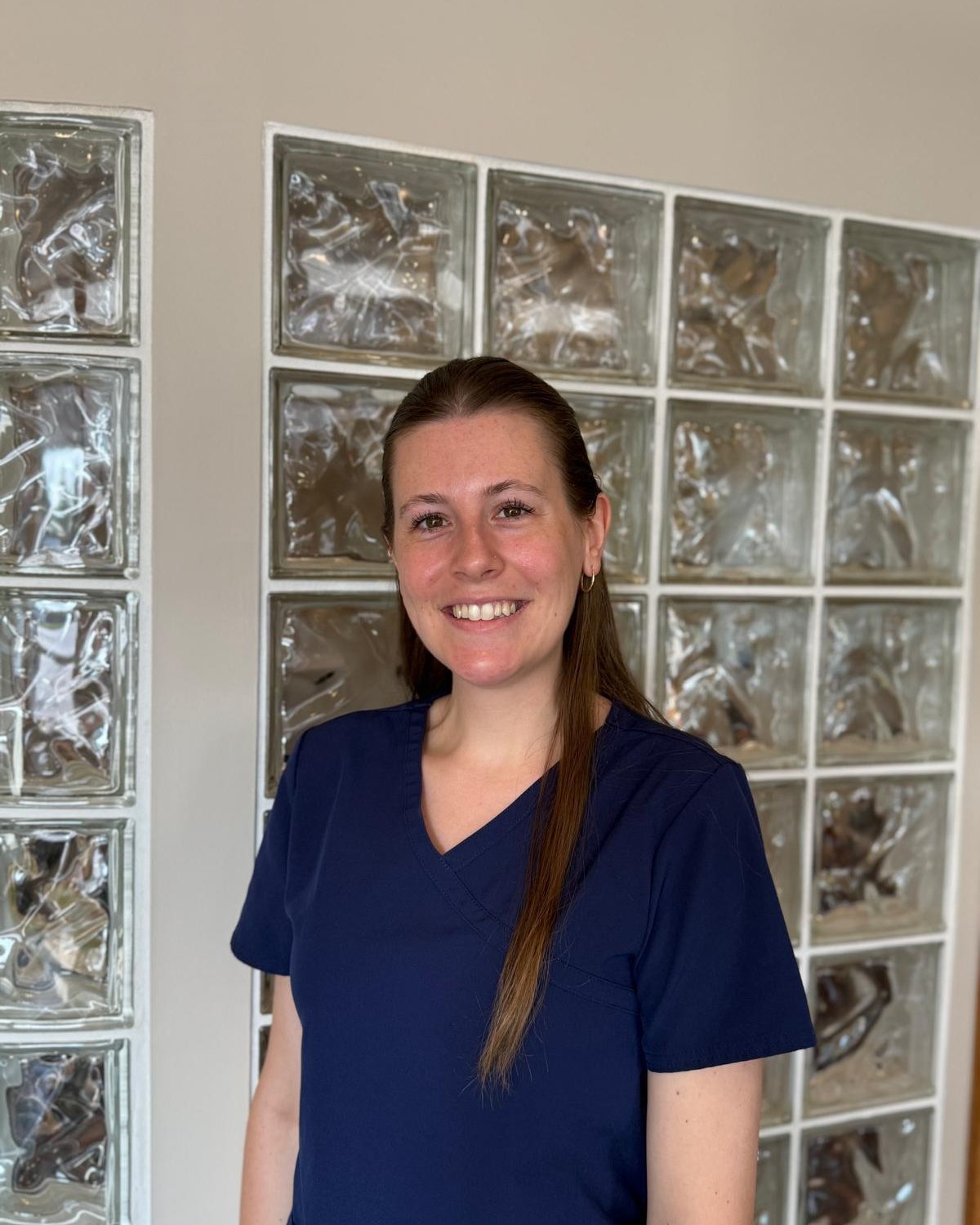 Carly, office manager, smiling in navy scrubs in front of a glass-block wall.