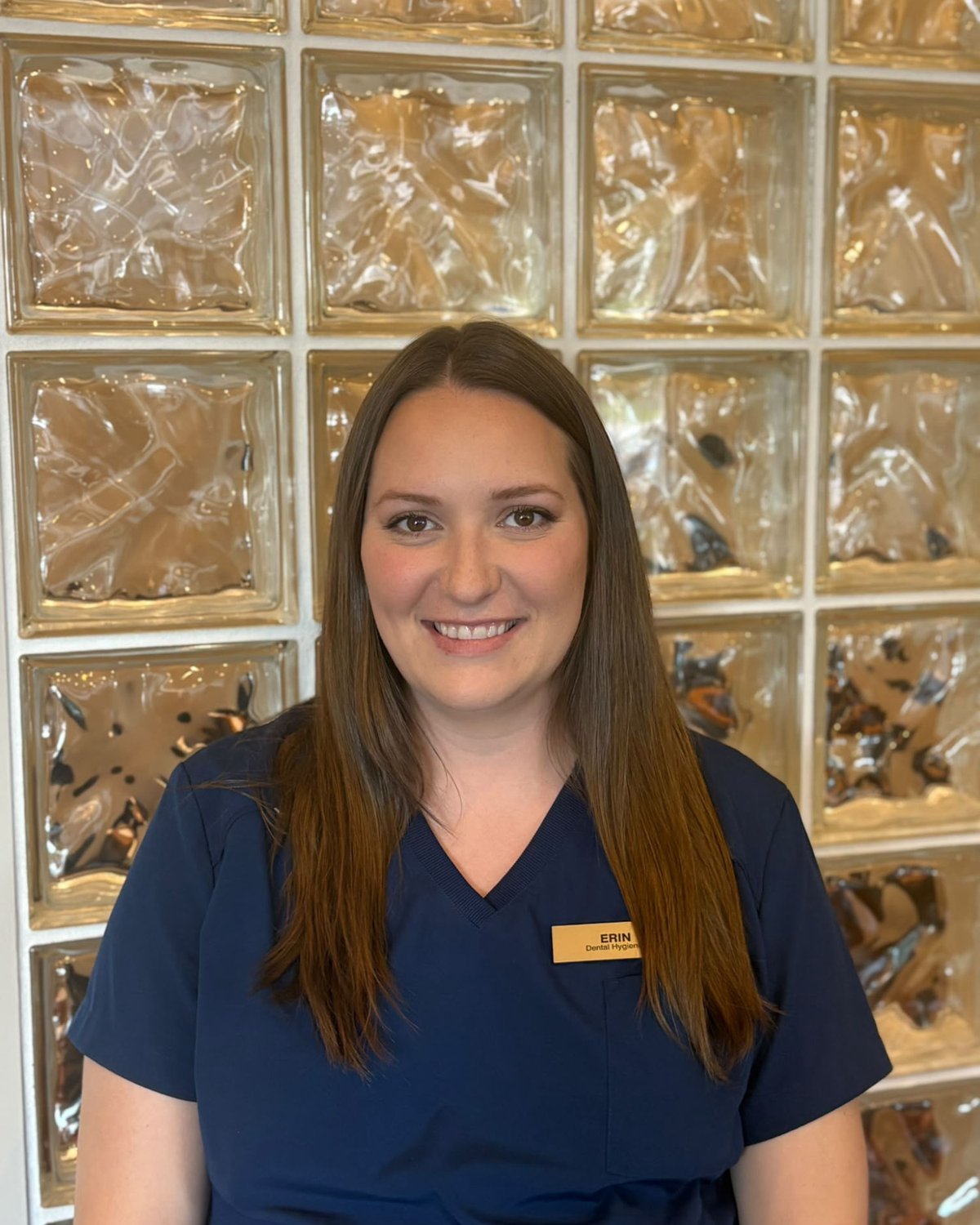 Erin, dental hygienist, smiling in navy scrubs in front of a glass-block wall.