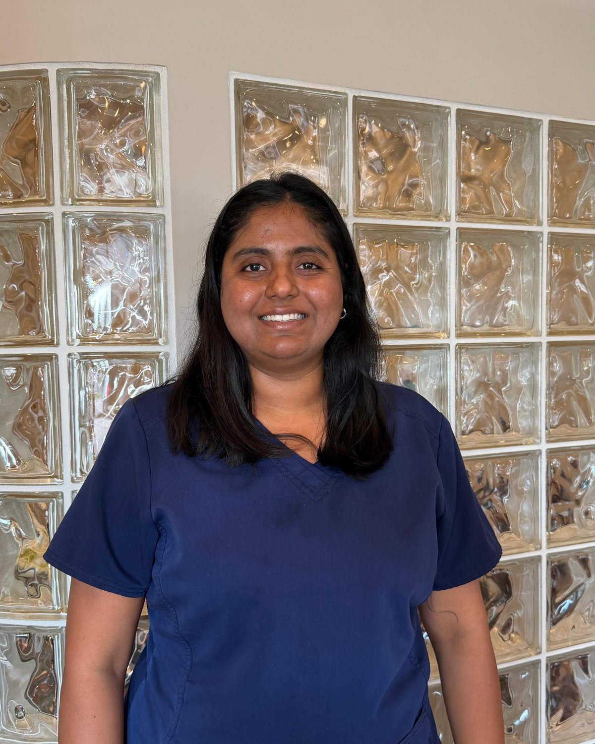 Prachi, front desk coordinator, smiling in navy scrubs in front of a glass-block wall.
