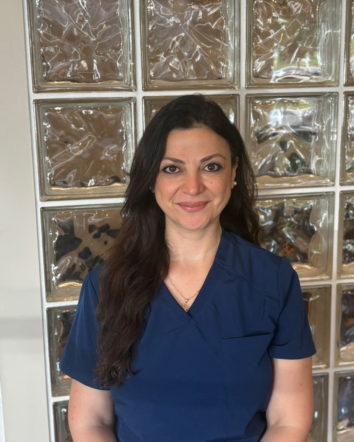 Ritta, dental assistant, smiling in navy scrubs in front of a glass-block wall.