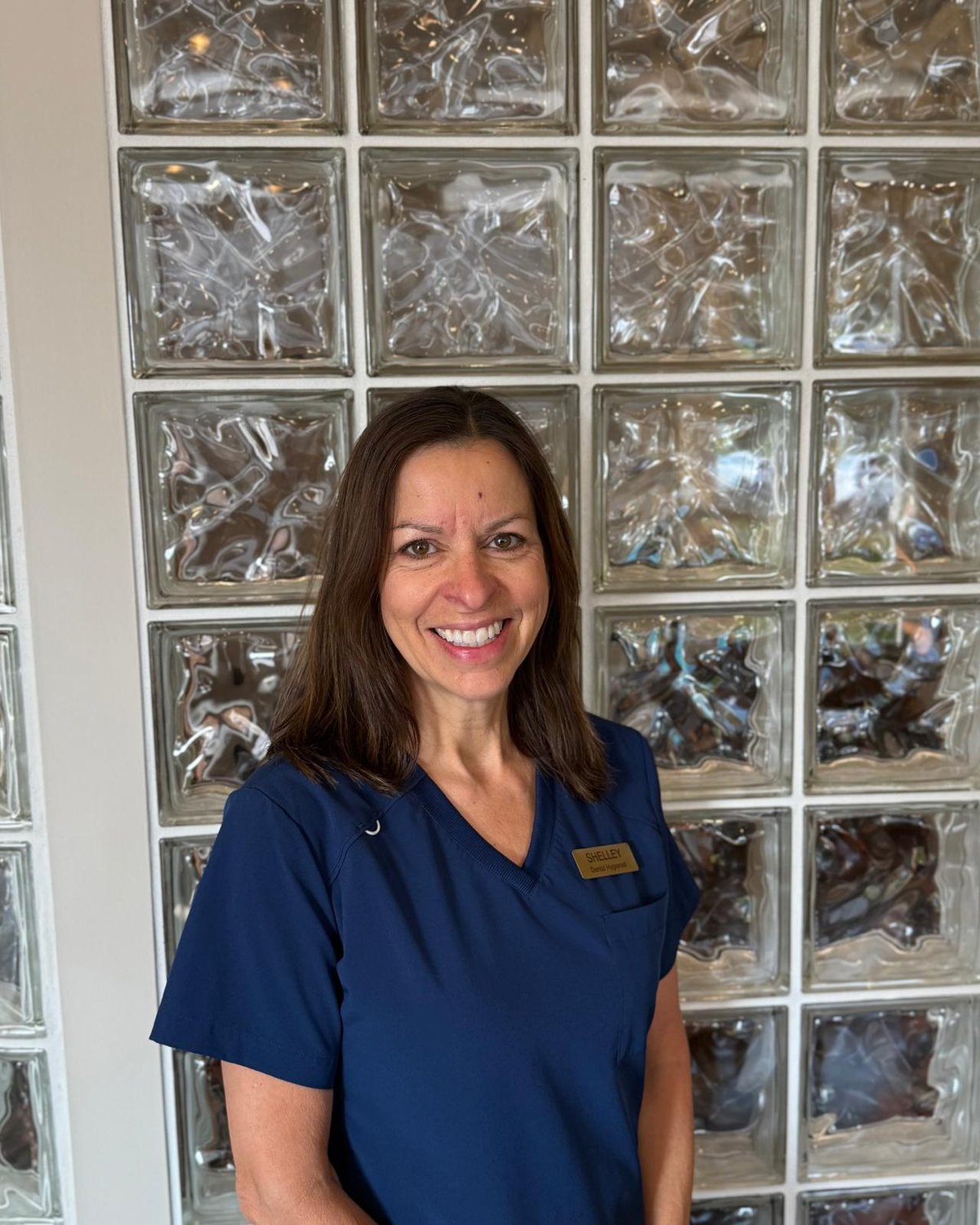 Shelley Karn, dental hygienist, smiling in navy scrubs in front of a glass-block wall.